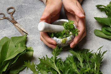 Woman grinding fresh green herbs in mortar at light grey table, closeup