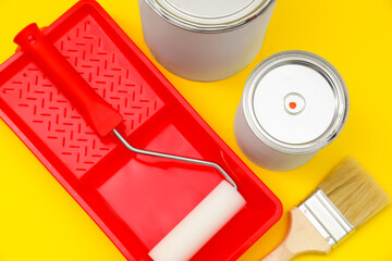 Cans of orange paint, brush, roller and container on yellow background, above view