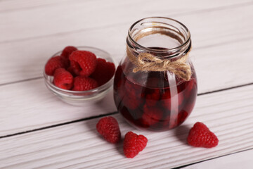 Jar of tasty canned raspberry jam and fresh berries in glass bowl on white wooden table