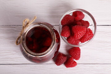 Jar of tasty canned raspberry jam and fresh berries on white wooden table, flat lay