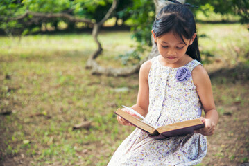 Asian Girl holding book reading at green park in natural garden. Young todler girl relaxation read open book self study. Happy child women smiling with happiness learning. Kid sit in green park