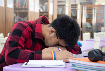  young asian gay wears rainbow wristband, sitting in library and taking a nap on table while doing his hard school project work, concept for raising teens and LGBT people daily activity.