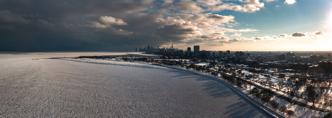 Aerial panorama of the Lake Michigan shoreline on the north side of Chicago covered in frozen ice 'pancakes' as clouds move past the downtown skyline in the distance and the sun begins to set.