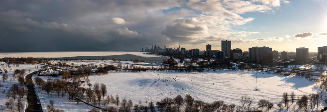 Beautiful winter aerial panorama photograph of people sledding in the snow on Cricket hill near Montrose Harbor with clouds over the downtown Chicago skyline in the distance at sunset.