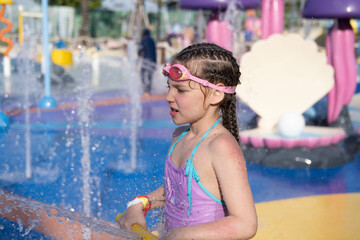 A child is playing in a water park. A little girl in a purple swimsuit and swimming goggles,...