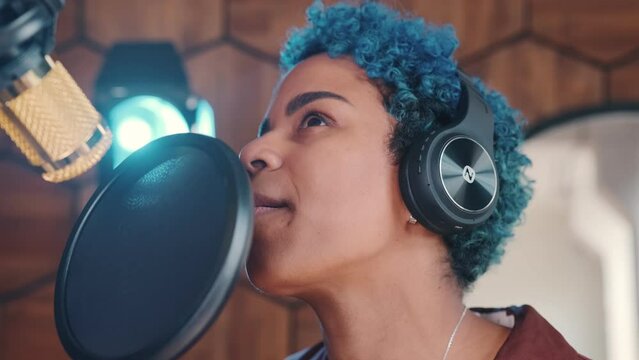 Young attractive African American woman singer uses noise-canceling microphone to record new POP or Hip Hop song in headphones to sell own songs on Internet stands in recording studio