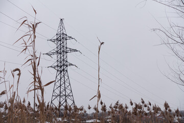 Support for high voltage transmission line. Metal structure on the outskirts of the village. Electric wires stretch along the village. Snow and dry reeds all around.