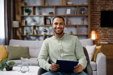 Young African American man sitting at home in a stylish office. Psychologist, coach or businessman looking at the camera and smiling. Beautiful teeth and smile.