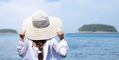 Fototapeta premium A Caucasian girl in a straw hat and a white tunic stands with her back to the camera and looks at the sea. Space for text.
