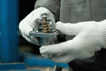 In the hands of an auto mechanic, a new thermostat for an automobile engine. Checking the integrity of a new spare part when replacing a faulty thermostat.Maintenance at a car service station.  - obrazy, fototapety, plakaty