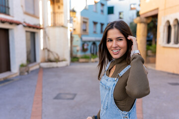 Happy young woman in denim overall standing on town street and fixing dark hair looking away during stroll