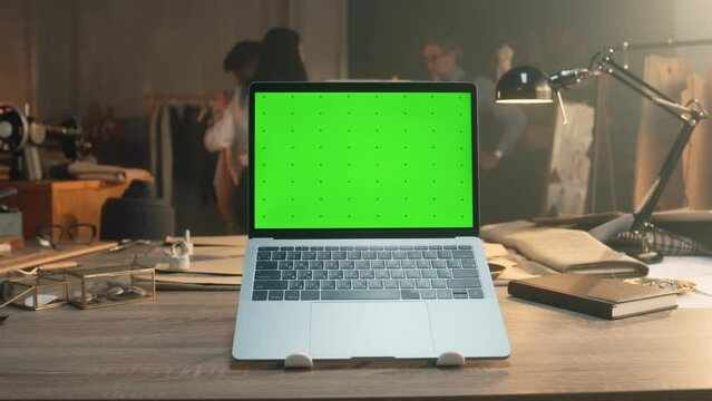 Cinematic Shot Of Laptop With Green Screen Standing On Table With In Tailoring Studio Or Luxury Atelier. Tailors Discussing With African American Client Business Suit On Background. Fashion Concept.