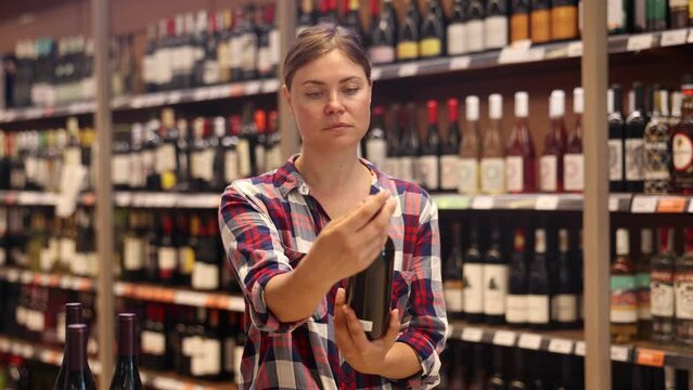 Young woman carefully reads the label on bottle while choosing wine in a supermarket. High quality 4k footage