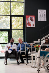 African american doctor holding clipboard explaining medical expertise to senior patient with wallking frame during consultation in hospital waiting area. Medicine service and concept