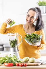 Smiling woman making spinach smoothie, putting leaves in blender.
