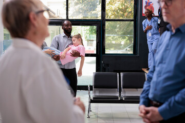 Worried dad entering in hospital waiting room holding unconscious little daughter in arms after fainting. Medical staff trying to help kid during checkup visit. Medicine service and concept