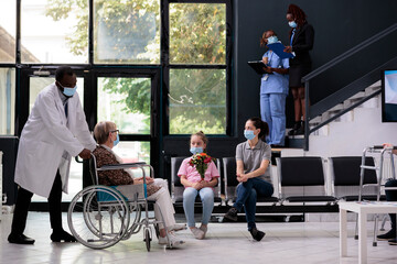 Physician doctor bringing grandmother in wheelchair to family after medical consultation during checkup visit appointment. Granddaughter holding bouqet flowers for senior woman. Virus epidemic