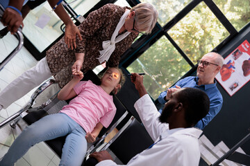 African american medic helping girl child after fainting on chair in hospital reception during...