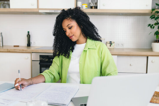 Horizontal Portrait Of Black Uni Student Girl In Casual Clothes With Long Curly Hair Sitting At Kitchen Table, Studying From Home, Holding Pen, Writing In Her Copybook. Online Education