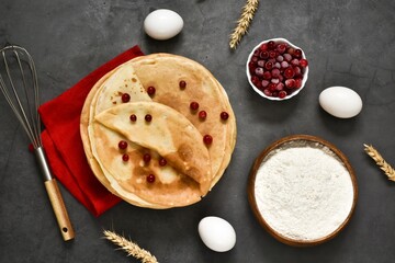 Pancakes on a plate with cranberries and ingredients for cooking on a gray concrete background. Traditional pancakes for Shrovetide. View from above. Copy space. Menu.