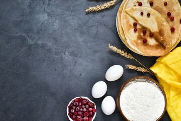 Pancakes on a plate with cranberries and ingredients for cooking on a gray concrete background. Traditional pancakes for Shrovetide. View from above. Copy space. Menu.