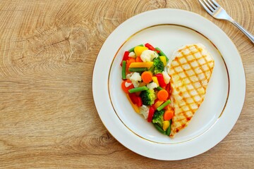 Heart shape grilled chicken breast with steamed vegetables on plate with wooden table background.Love healthy food concept for Valentine's day and Mother's day.Top view.Copy space