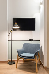 Closeup of modern comfortable armchair with dark blue upholstery bright ceiling lights and metal floor lamp in room with white walls and wooden parquet. Behind TV on wall for watching shows.
