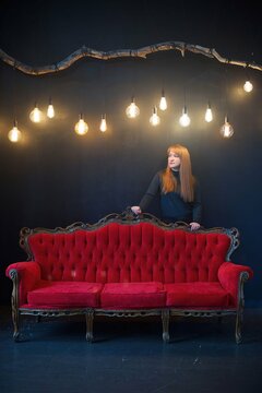 A Woman Is Standing In A Dark Room Behind A Red Sofa. Light Bulbs Hang From Above As Lighting.