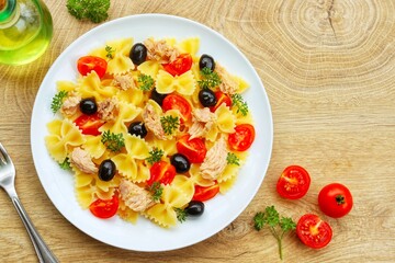 Farfalle pasta salad with canned tuna in olive oil, cherry tomatoes,black olives,parleys,olive oil and peppers on plate with wooden background.Healthy Italian summer salad.Top view.Copy space

