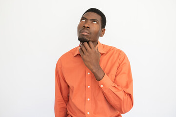 Portrait of pensive young man rubbing chin over white background. African American guy wearing orange T-shirt looking up with thoughtful expression. Thinking and planning concept