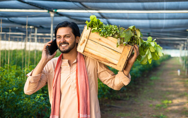 tracking shot happy farmer talking on mobile phone while carrying basket of vegetables at...