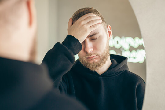 Sad young man with a beard in front of a mirror, mental health concept.