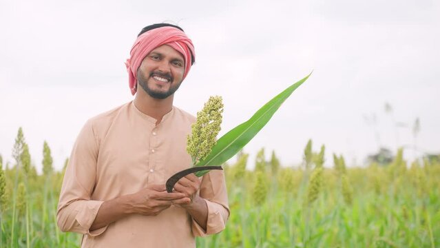 Happy smiling young farmer holding crop and sickle by looking camera at corn or maize field - concept of real rural people, happiness and cultivation