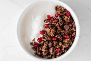 Bowl of greek yogurt with chocolate oat granola and pomegranate seeds on white marble table. Top view