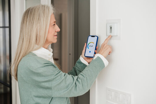 Mature Woman Adjusting Thermostat With Smart Phone At Home