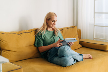 Attractive middle-aged caucasian woman sitting on a sofa works on a computer tablet.