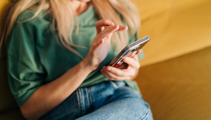 Unrecognizable woman browsing media on mobile phone while sitting on yellow sofa at home.