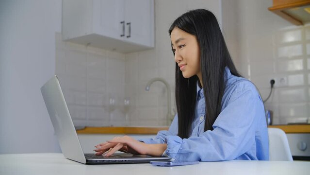 Asian BIPOC female typing text on laptop keyboard in home kitchen. Freelancer POC works on modern notebook pc. Vietnamese girl using computer with a smile.