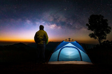 A glowing blue camping tent man standing in a high place looking up in wonder at the Milky Way...