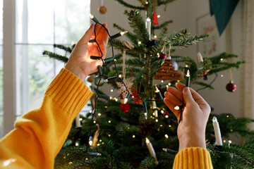 Hands of woman putting illuminated string lights on Christmas tree at home