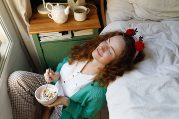 Smiling woman with eyes closed holding cereal bowl by bed at home