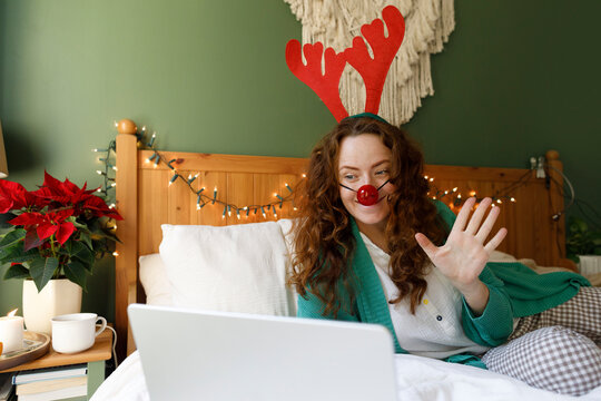 Woman wearing antler and clown's nose waving on video call through laptop in bedroom