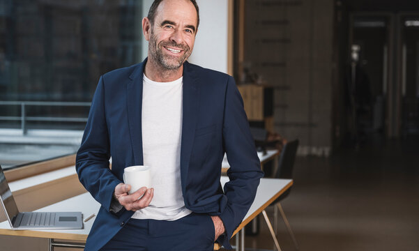 Happy businessman with coffee cup leaning on desk in office