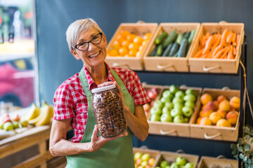 Woman works in fruits and vegetables shop. She is holding jar with almond.