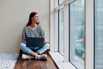 Business woman sitting with a laptop at the window full-length and smiling, happy woman work online in the office, copy space