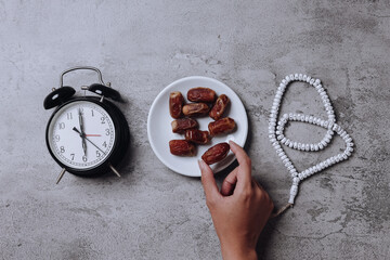Hand take date fruit on hand on gray background, arranged with alarm clock showing iftar time and prayer beads. 