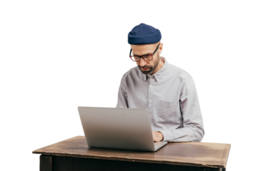Fashionable male student types publication on laptop computer, prepares for exam, spends time on education and getting knowledge, poses at old desk, wears stylish hat, glasses and white shirt