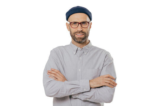 Horizontal shot of satisfied self confident male designer wears stylish headgear, dressed in white shirt, keeps arms folded, has dark stubble, isolated over white background. People and style concept
