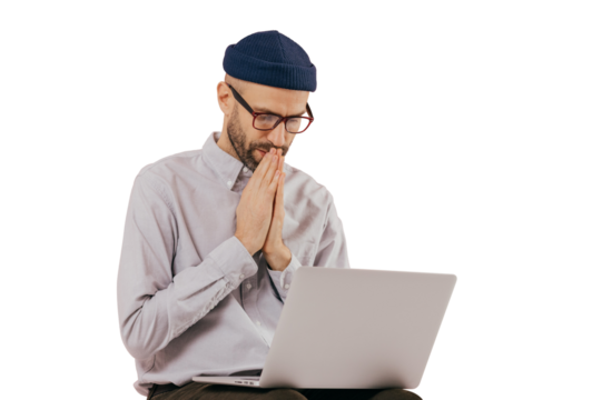 Professional male programmer keeps hans in praying gesture, looks at screen of laptop computer, believes in successful result of his work, wears formal shirt and hat, isolated over background