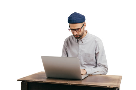 Fashionable male student types publication on laptop computer, prepares for exam, spends time on education and getting knowledge, poses at old desk, wears stylish hat, glasses and white shirt
